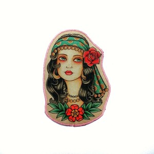 May include: A colorful patch featuring a woman with dark hair, green eyes, and red lips. She wears a green headscarf with gold dots and a red rose. A red flower is at the bottom. The patch has a pink border.