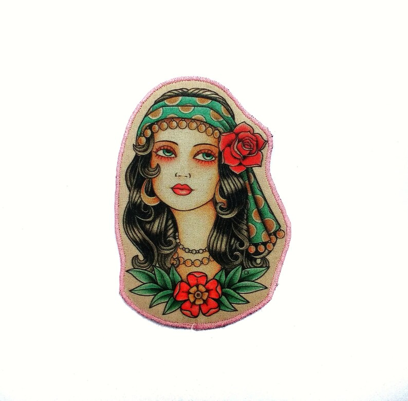 May include: A colorful patch featuring a woman with dark hair, green eyes, and red lips. She wears a green headscarf with gold dots and a red rose. A red flower is at the bottom. The patch has a pink border.
