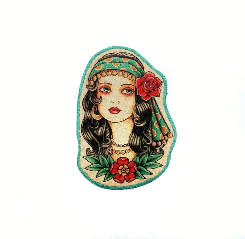 May include: A colorful patch featuring a woman with dark hair, wearing a green headscarf with gold polka dots and a red rose. The woman has a necklace and a red flower at the bottom. The patch has a teal border.