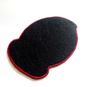 May include: A black felt patch with a red stitched border. The patch has a unique, rounded shape and is set against a white background. The texture of the felt is visible, adding depth to the design.