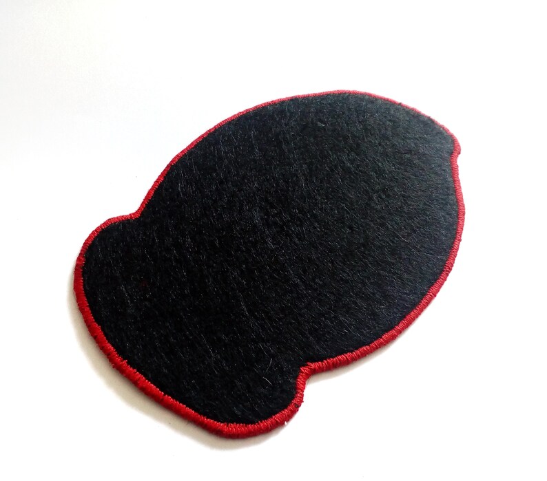 May include: A black felt patch with a red stitched border. The patch has a unique, rounded shape and is set against a white background. The texture of the felt is visible, adding depth to the design.