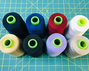 May include: Eight spools of thread in various colors, including beige, black, blue, red, white, and purple, are arranged in a circle on a green cutting mat.