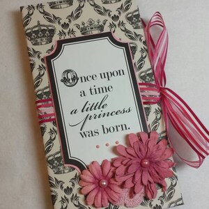 May include: A decorative journal with a cream and black crown pattern. The journal features a white label with the text "Once upon a time a little princess was born." Pink paper flowers and a pink ribbon add a decorative touch.