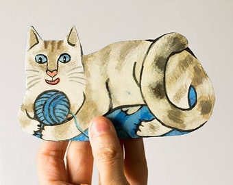May include: A hand holds a painted wooden cutout of a white cat with blue eyes playing with a ball of yarn. The cat is lying down with its tail curled around its body.
