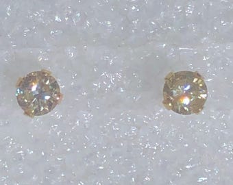 May include: A pair of round-cut stud earrings featuring clear, faceted stones set in gold-toned metal prongs. Each earring displays a brilliant, reflective surface with subtle light refractions, showcasing a classic circular design held securely by four small metal claws.