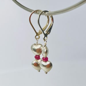 May include: Silver-toned earrings with heart-shaped charms and small red beads. The earrings are suspended from lever-back closures. The design features a combination of polished silver and vibrant red accents, creating a romantic and elegant look.