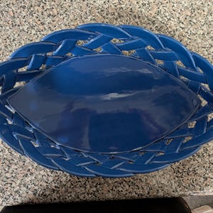 May include: A glossy, deep blue ceramic basket with an elongated, eye-shaped center and a decorative woven lattice border. The piece features a smooth, solid-colored base and a structured, interlaced rim design in a matching dark blue finish.