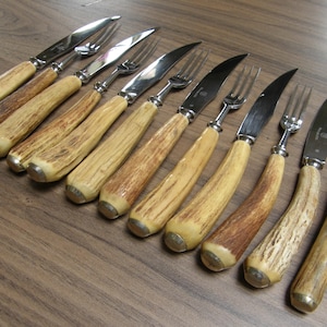May include: A set of antique cutlery, including knives and forks, with natural-toned antler handles and stainless steel blades. The set is arranged on a wooden surface, showcasing the rustic elegance of the dining set.