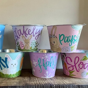 May include: A collection of hand-painted metal buckets in pastel colors, featuring Easter-themed designs. Each bucket has a different name written on it, along with illustrations of bunnies, eggs, and floral accents. The buckets are displayed on a wooden surface.