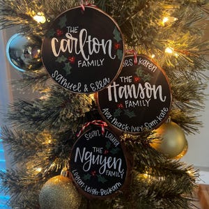 May include: Three round, black wooden Christmas ornaments with white lettering and red accents hang on a Christmas tree. The ornaments display family names: "The Carlton Family", "The Hanson Family", and "The Nguyen Family".
