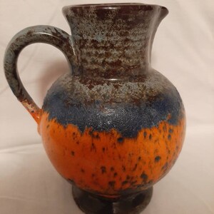 Könnte beinhalten: Ein großer orangefarbener, blauer und brauner Keramik Krug mit einer breiten Öffnung und einem Henkel. Der Krug hat eine gefleckte Glasur und ein rustikales Aussehen.