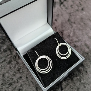 May include: Silver triple hoop earrings displayed in a gray jewelry box with a black velvet interior. The earrings have a simple, modern design, with three graduated hoops suspended from a hook.