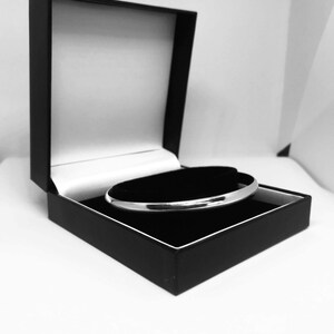 May include: A silver bangle bracelet presented in an open black jewellery box with a black velvet interior. The bracelet sits on a curved black platform. The background is white.