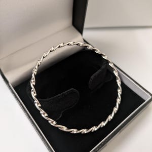 May include: A silver bangle with a twisted rope design, displayed in a black jewellery box with a black velvet interior. The bracelet is circular and appears to be made of metal.
