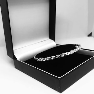 May include: A silver bangle bracelet with a braided design sits in a black velvet jewellery box.