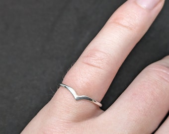 May include: A silver-colored ring with a V-shaped design. The ring is worn on a finger, with a dark background. The ring's minimalist design makes it suitable for everyday wear.