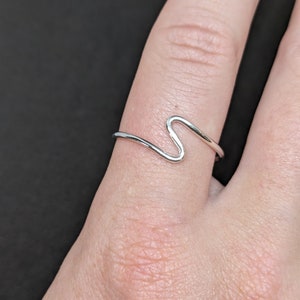 Könnte beinhalten: Ein silberner Ring mit einem wellenförmigen Design. Der Ring wird an einem Finger getragen.