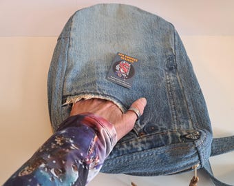 May include: A denim bag made from recycled jeans. The bag is light blue with visible stitching and a zipper closure. A small tag with the text "REN SQUIRES DESIGNS" is attached to the bag.