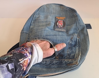 May include: A denim backpack with a rounded shape and a small pocket. The backpack has a faded blue color and a small tag that reads "REN SQUIRES DESIGNS". A hand is visible in the image.