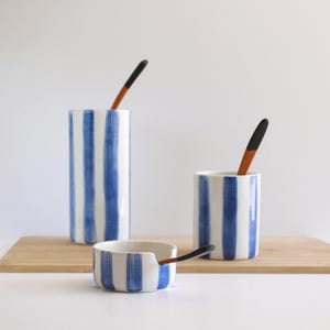 Può includere: Tre pezzi di ceramica con strisce verticali blu e bianche. Un cilindro alto, un cilindro più corto e una piccola ciotola, ciascuno con un cucchiaio di legno. Gli oggetti sono su un tagliere di legno.