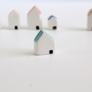 May include: A selection of miniature ceramic houses with white walls and colourful roofs. The houses have small black square windows. The roofs are painted in various colours, including blue, pink, and green. The houses are arranged on a white surface.