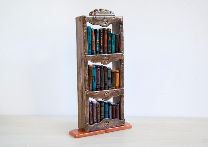 May include: A decorative ceramic bookshelf with three shelves, filled with miniature books. The bookshelf is brown with ornate detailing, and the books have colorful spines. The base is a reddish-brown color.
