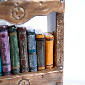 May include: A miniature bookshelf with a collection of colorful, handcrafted books. The books have various hues, including blue, purple, green, and orange, and are arranged on a brown wooden shelf. The shelf has decorative metal accents.