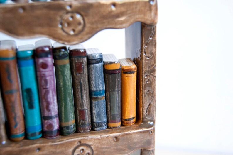 May include: A miniature bookshelf with a collection of colorful, handcrafted books. The books have various hues, including blue, purple, green, and orange, and are arranged on a brown wooden shelf. The shelf has decorative metal accents.