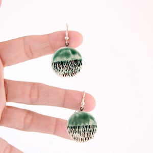 Puede incluir: Un par de pendientes colgantes redondos con un esmalte verde brillante en la mitad superior y un patrón texturizado de rayas verticales en marrón oscuro y blanco en la mitad inferior, suspendidos de ganchos de metal plateado.