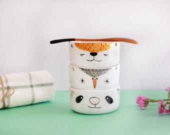 May include: A stack of three white ceramic bowls, each adorned with a different animal face: a fox, a bird, and a panda. A wooden spoon rests on top. A rolled-up towel and pink flowers are in the background.