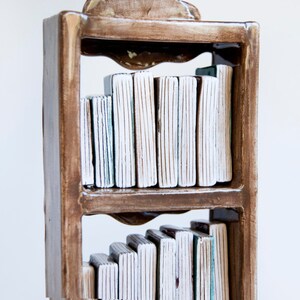 May include: A handcrafted ceramic bookshelf sculpture with two shelves. The frame is a warm brown, and the books are white with textured vertical lines. The sculpture has a decorative top and is set against a white background.
