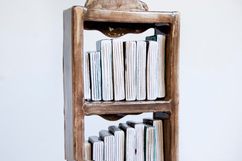 May include: A handcrafted ceramic bookshelf sculpture with two shelves. The frame is a warm brown, and the books are white with textured vertical lines. The sculpture has a decorative top and is set against a white background.