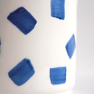May include: A white ceramic vase with a pattern of blue brushstrokes. The blue shapes are rectangular and irregularly shaped, creating a modern, artistic design. The vase is cylindrical and appears to be handmade.