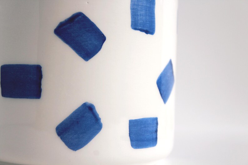 May include: A white ceramic vase with a pattern of blue brushstrokes. The blue shapes are rectangular and irregularly shaped, creating a modern, artistic design. The vase is cylindrical and appears to be handmade.
