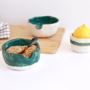 Può includere: Ciotole in ceramica fatte a mano in bianco e verde acqua. Una ciotola contiene semi con un cucchiaio di legno. Un'altra ciotola contiene un limone. Le ciotole sono su un tagliere di legno e una superficie bianca. Un panno a scacchi è sullo sfondo.