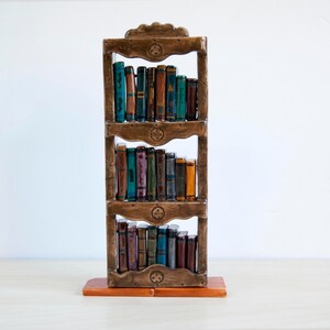 May include: A decorative wooden bookshelf with three shelves, filled with miniature, colorful books. The bookshelf is a warm brown color, with carved details and a rectangular base. The books have various colors and designs, adding a whimsical touch to the piece.