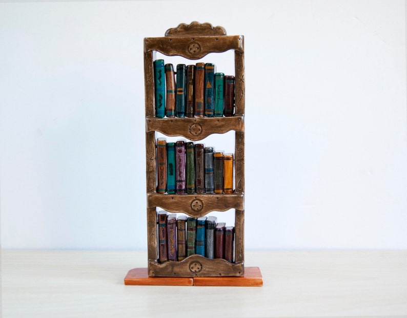 May include: A decorative wooden bookshelf with three shelves, filled with miniature, colorful books. The bookshelf is a warm brown color, with carved details and a rectangular base. The books have various colors and designs, adding a whimsical touch to the piece.