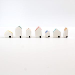 Può includere: Una collezione di sette casette in ceramica in miniatura con pareti bianche e tetti colorati. Ogni casa ha una porta nera. I tetti sono gialli, verdi, rossi, blu e beige. Le case sono disposte in fila su una superficie bianca.