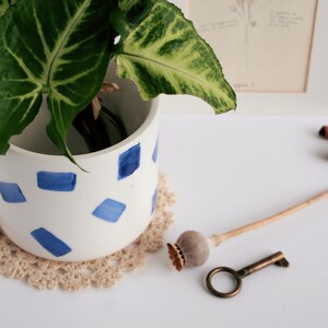 May include: A white ceramic planter with blue geometric shapes holds a green plant with large leaves. A beige crocheted doily sits beneath the planter. A vintage key, a wooden object, and two cinnamon sticks are also on the white surface.