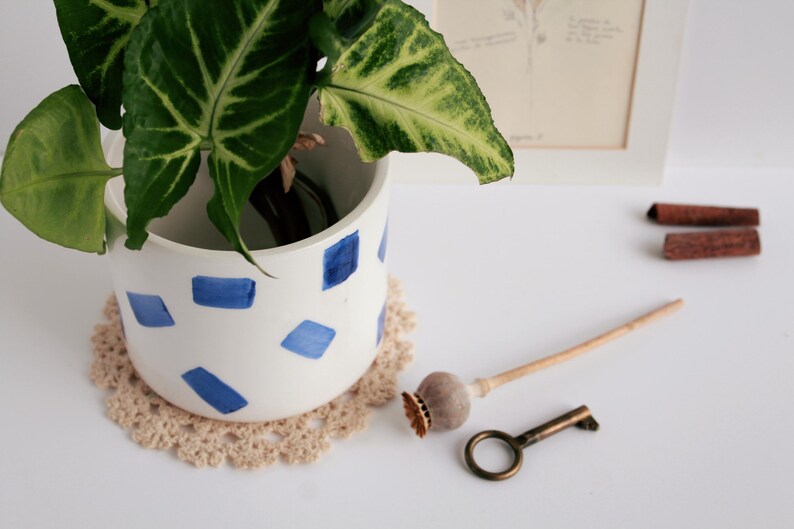 May include: A white ceramic planter with blue geometric shapes holds a green plant with large leaves. A beige crocheted doily sits beneath the planter. A vintage key, a wooden object, and two cinnamon sticks are also on the white surface.