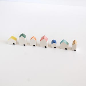Pode incluir: Uma coleção de pequenas figuras de casas de cerâmica com paredes brancas e telhados coloridos. Os telhados são pintados em amarelo, verde, laranja, vermelho, azul e bege. Cada casa tem uma pequena porta quadrada preta.