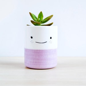 Può includere: Un piccolo vaso di ceramica bianca con una base color lavanda e un design a faccina sorridente. Il vaso contiene una pianta succulenta verde. Il design presenta occhi neri e lentiggini. Il vaso è su una superficie di legno chiaro.