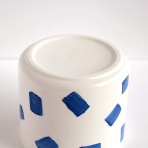 May include: White ceramic mug with a cylindrical shape, decorated with irregular blue brushstrokes. The mug has a smooth, glossy finish and a simple, modern design. The bottom of the mug is visible in the image.