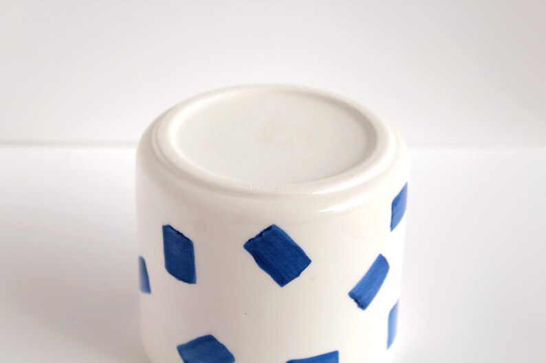 May include: White ceramic mug with a cylindrical shape, decorated with irregular blue brushstrokes. The mug has a smooth, glossy finish and a simple, modern design. The bottom of the mug is visible in the image.