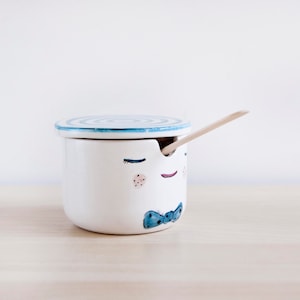 Op de afbeelding: Een witte keramische suikerpot met een deksel en een houten lepel. De pot heeft een grillig gezichtsontwerp met gesloten ogen, roze wangen en een blauwe vlinderdas. Het deksel heeft een blauw spiraalontwerp.
