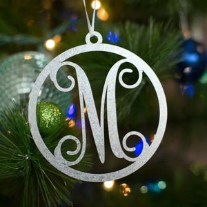 May include: Silver Christmas ornament with a decorative letter M in a circle. The ornament is hanging on a green Christmas tree.
