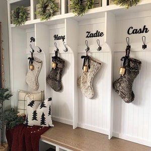 May include: A white storage unit with four hanging stockings, each adorned with a bell and black ribbon. The unit features name labels and decorative wreaths. A bench below holds pillows and a red blanket, creating a festive holiday scene.