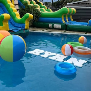 May include: A blue pool with a white inflatable slide, a yellow and green inflatable bounce house, and a white inflatable name spelling "RYAN". There are also several inflatable pool toys, including a blue and green beach ball, a red and white beach ball, and a yellow beach ball.