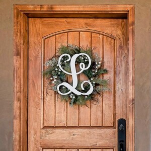 May include: A wooden front door with a wreath featuring a white letter L on a brown background. The door has a black door handle and a window pane to the right.