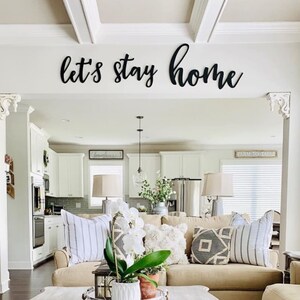 Puede incluir: Una sala de estar con un sof&aacute; beige y una mesa de centro. El sof&aacute; tiene varios cojines, incluyendo un coj&iacute;n geom&eacute;trico en blanco y negro. La mesa de centro tiene una bandeja blanca con una orqu&iacute;dea en maceta y otras plantas. La pared sobre el sof&aacute; tiene un letrero de madera negro que dice "let's stay home".